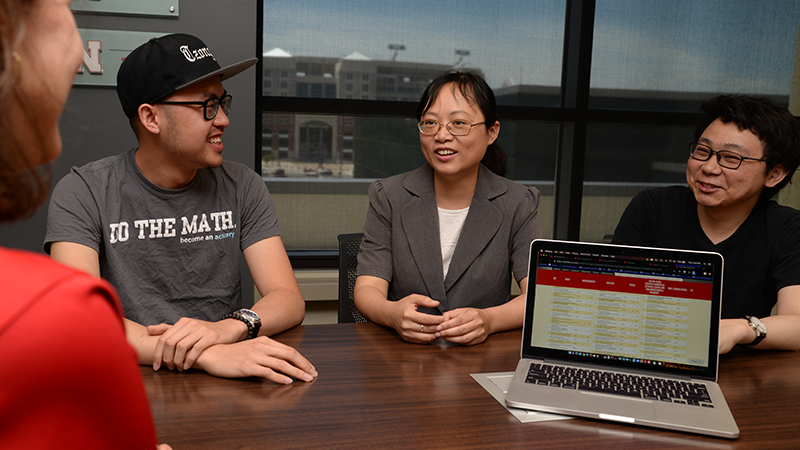 Group seated at a table, laptop displaying a red website, window view of buildings behind.