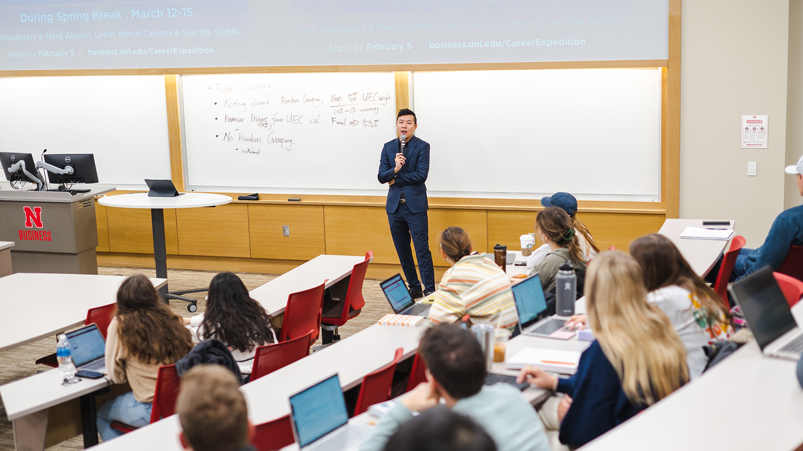 Professor speaking to students in a classroom with whiteboards and red seating.