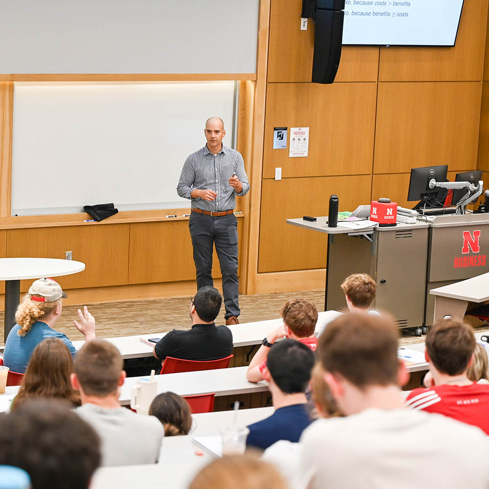 Speaker addressing students in a classroom with wood-paneled walls and a whiteboard.