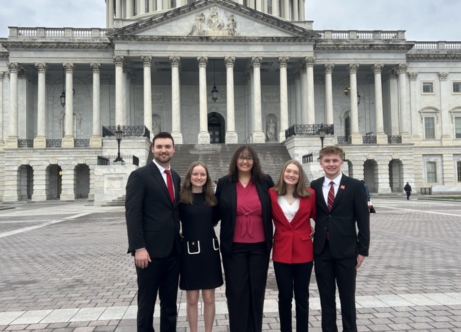 Five people in formal attire standing outside a large white building with columns and steps.