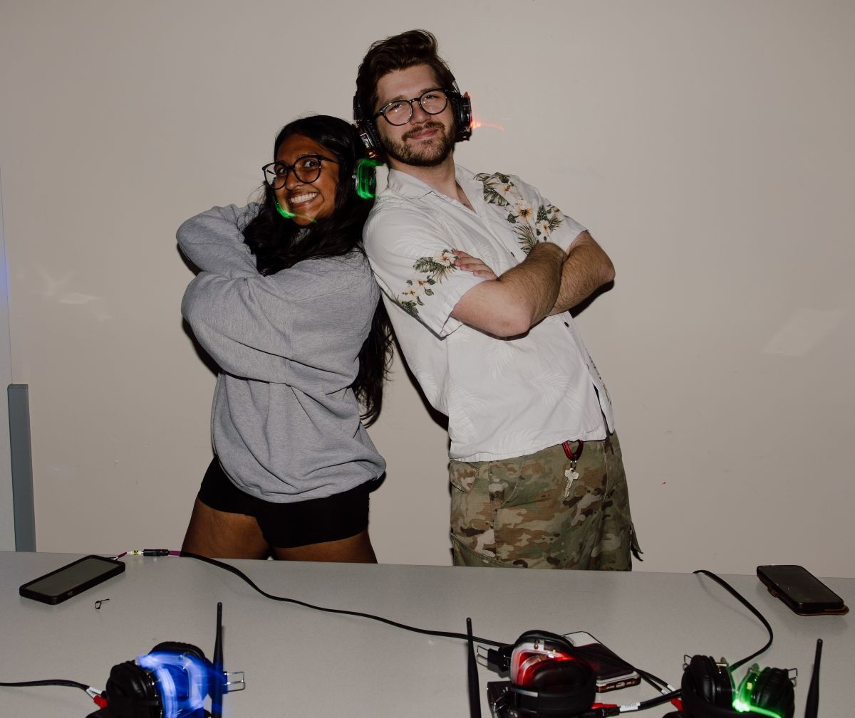Two people smiling with arms crossed, wearing headphones with glowing lights, standing by a table.