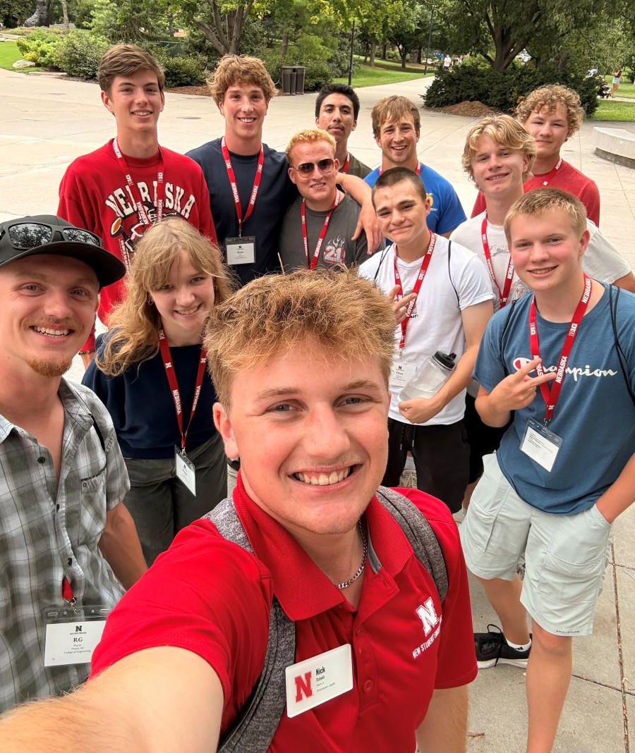 Group of smiling young adults wearing name tags and lanyards outdoors on a sunny day.