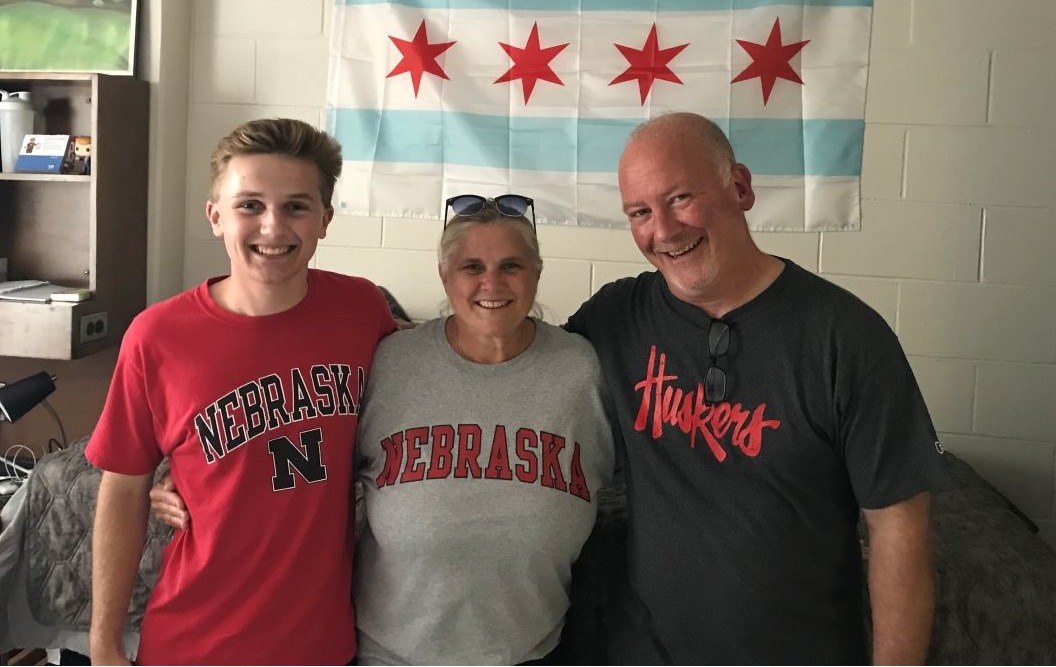 Nick and his parents wearing Nebraska shirts, with a Chicago flag on the wall behind them.