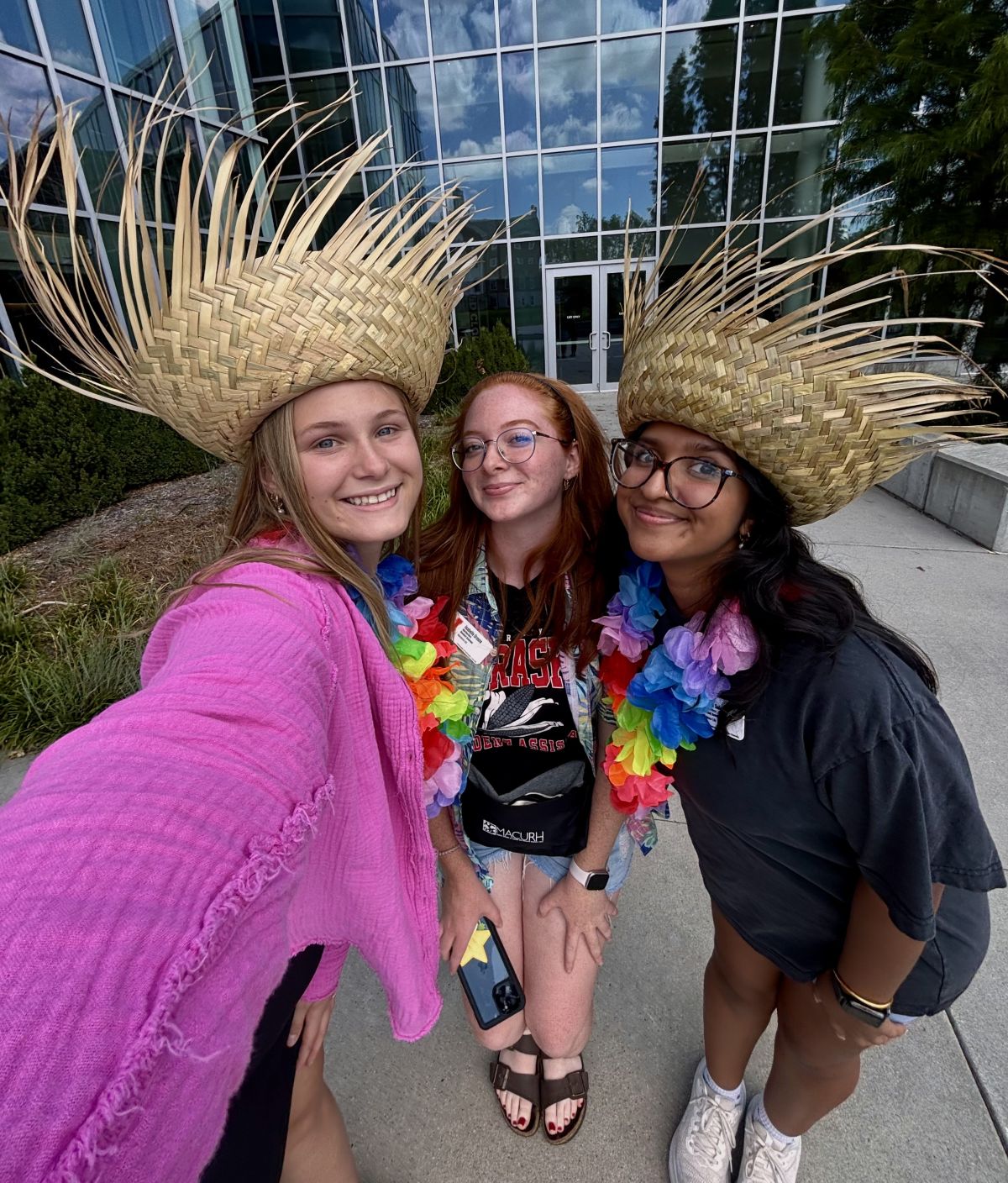 Subedi and friends wearing straw hats and colorful leis, standing outdoors near a glass building.