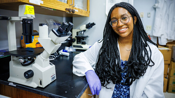 Person in lab coat and gloves near a microscope in a laboratory setting.