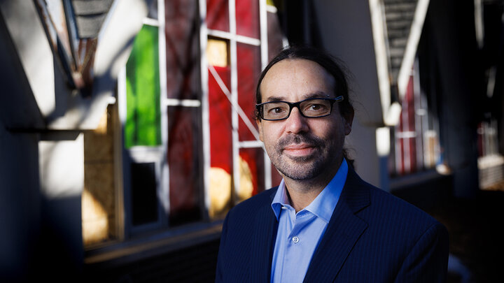 Philip Schwadel is photographed in front of a stained-glass window, in shadows.