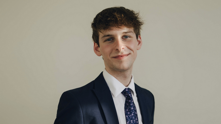 Luke McDermott, who is white and has brown hair, smiles in a blue suit and tie.
